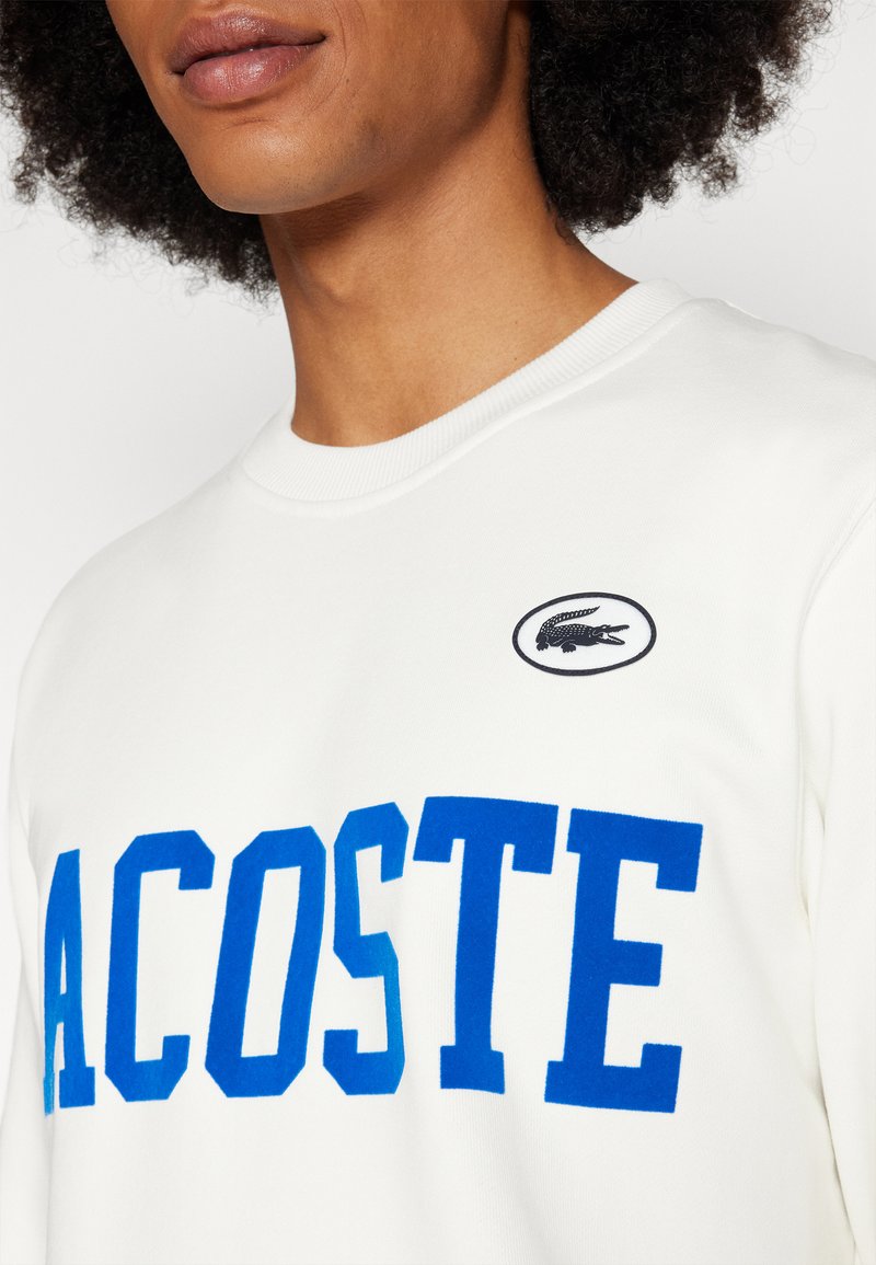 Felpa bianca con grande scritta blu "LACOSTE" e un piccolo logo ricamato nero sul petto. Collo rotondo e polsini a coste.
