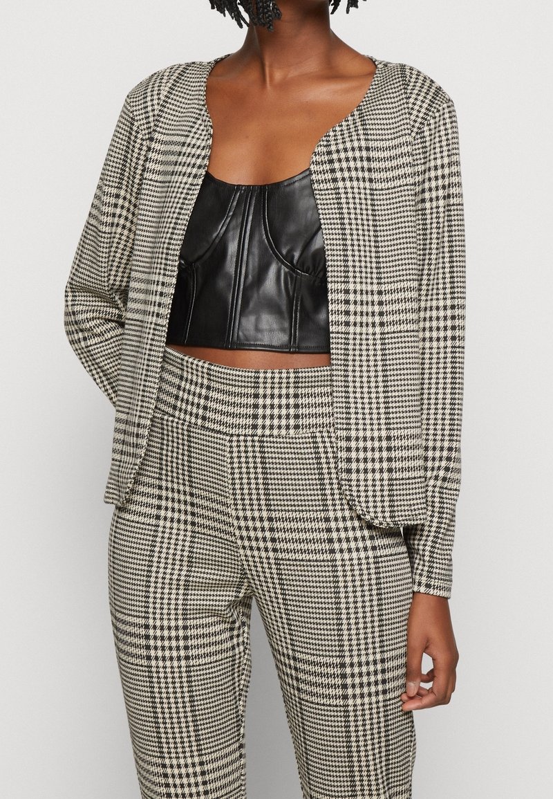 Femme portant un crop top en cuir noir sous un blazer à motif pied-de-poule noir et blanc et un pantalon assorti.