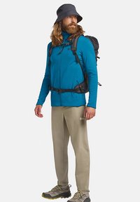 Giacca blu con cerniera, pantaloni leggeri color sabbia, zaino nero, scarpe da trekking grigie con accenti gialli e cappello a tesa larga nero.