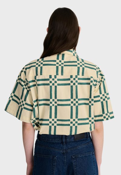 Personne aux longs cheveux foncés portant une chemise beige à manches courtes avec des motifs en grille verte et un jean bleu foncé, vue de dos.