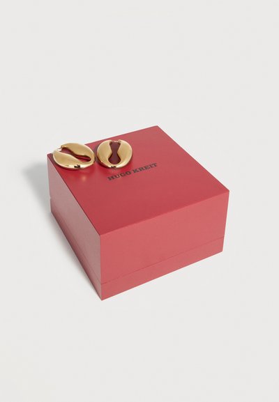 Guld ovalformede øreringe med åbent center placeret på en lukket rød Hugo Kreit smykkeskrin mod en hvid baggrund.