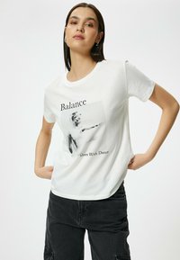 Hvid bomulds T-shirt med et sort-hvidt kunstnerisk grafisk print med teksten "Balance møder dans" centreret nedenfor.