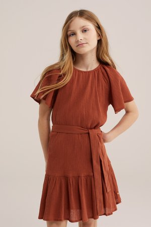 Ung pige med langt blondt hår iført en rustfarvet kortærmet kjole med flæset kant og bundet taljebælte, stående mod en ensfarvet baggrund.