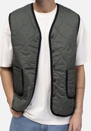 Person iført en ærmeløs mørkegrøn quiltet vest med sort kant over en hvid kortærmet skjorte og blå jeans.