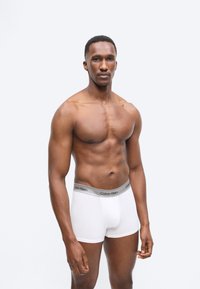 Homme musclé aux cheveux courts portant un boxer blanc Calvin Klein, debout face à un fond gris clair uni, regardant droit devant lui.