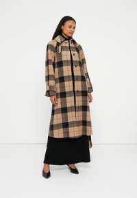 Long manteau à carreaux dans des tons beige et noir. Fabriqué en laine, il présente un col, une fermeture à boutons et des fentes latérales pour un mouvement facile.