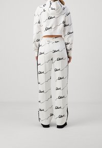 Femme portant un pantalon blanc ample et un crop hoodie assorti avec un motif script noir "Colucci" répété, vue de dos.