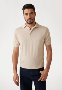 Bežs īsām piedurknēm polo krekls ar klasisku apkakli, ar gludu tekstūru un divu pogu aizdari, savienots ar tumši džinsu biksēm.