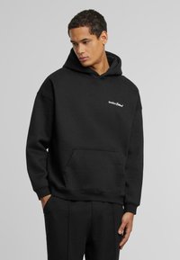 Sudadera de algodón negra con un bolsillo canguro frontal, puños y dobladillo acanalados, y un logo blanco en el lado izquierdo del pecho. Diseño simple y de corte relajado.