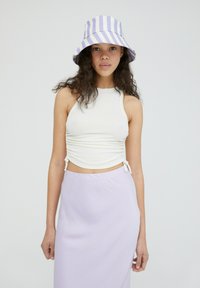 Wit geribbelde crop top met zijkanten, gecombineerd met een lavendelkleurige maxi rok. Het model draagt een gestreepte paarse en witte bucket hat.