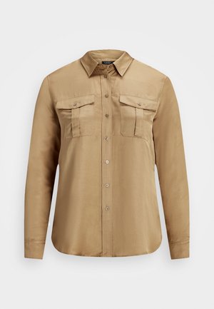 Chemise à manches longues de couleur beige, boutonnée, avec deux poches poitrine boutonnées et un col pointu, présentée sur un fond blanc.