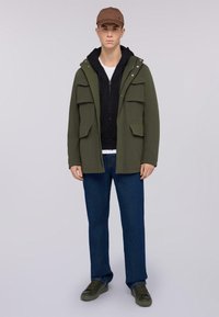 Parka verde oliva con cuatro bolsillos frontales, superpuesta a una sudadera negra, combinada con vaqueros azules y zapatillas verde oscuro. Modelo llevando una gorra marrón.