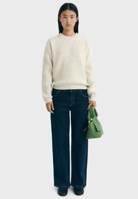 Junge Frau mit glattem schwarzen Haar, die einen cremefarbenen Strickpullover, dunkelblaue weite Jeans und schwarze Schuhe trägt und eine grüne Handtasche an ihrer Seite hält.