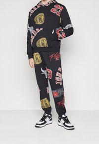 Sudadera con capucha y pantalones deportivos negros con varios estampados gráficos en rojo y dorado, que incluyen logotipos y texto, combinados con zapatillas negras y blancas.