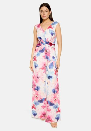 Femme debout portant une combinaison sans manches à col en V avec un pantalon à jambes larges dans un motif floral rose, bleu et blanc sur un fond uni.