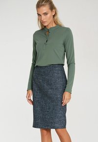 Zielona bluzka z długim rękawem i dekoltem w kształcie łezki, w połączeniu z szaro teksturowaną spódnicą o długości do kolan.