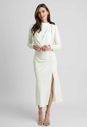 Kobieta z długimi brązowymi włosami, ubrana w białą, długorękawową sukienkę do połowy łydki z marszczeniem po bokach i rozcięciem, stoi i się uśmiecha, widoczne beżowe szpilki.