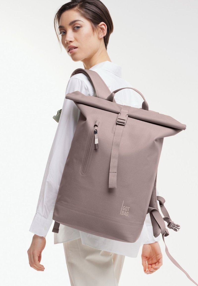 Sac à dos à rabat en taupe clair, fabriqué à partir de matériau durable. Comprend une poche zippée, des bretelles réglables et un détail de logo à l'avant.