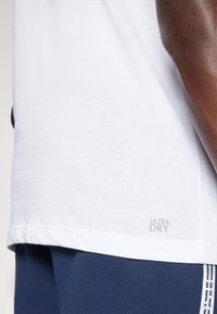 Biała koszulka z krótkim rękawem wykonana z miękkiego materiału, z szarym napisem "ULTRA DRY" nadrukowanym w pobliżu dolnego brzegu. W połączeniu z granatowymi spodniami.