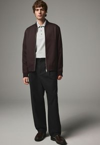 Jachetă bomber de un burgund închis peste un tricou polo de culoare gri deschis, cu pantaloni negri și pantofi loafers maro. Material neted cu un design simplu și modern.