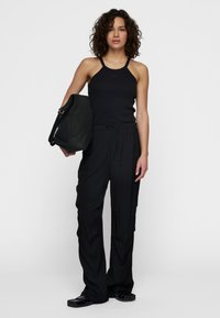 Zwarte mouwloze geribbelde top, ontspannen zwarte broek met koord, en een zachte zwarte tas, allemaal in een matte afwerking. Model staat tegen een effen achtergrond.