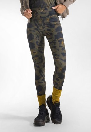 Leggings med grønt og sort camouflagemønster båret med gule sokker, sorte chunky sneakers og en brun jakke.