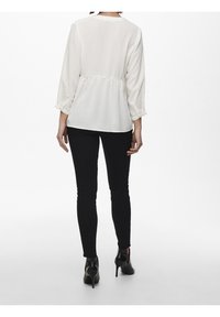 Blusa bianca a pieghe con maniche a tre quarti, dettaglio della cintura in vita e scollatura arrotondata, abbinata a pantaloni neri aderenti e scarpe con tacco alto.