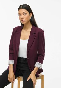 O tânără cu păr lung și închis la culoare stă pe un taburet din lemn, purtând un sacou violet cu manșete în dungi, un top alb și pantaloni negri, privindu-ne în cameră.