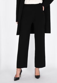 Zwarte, wijde broek met een gladde textuur en een hoge taille, gecombineerd met zwarte hoge hakken. Een minimalistische, moderne uitstraling.