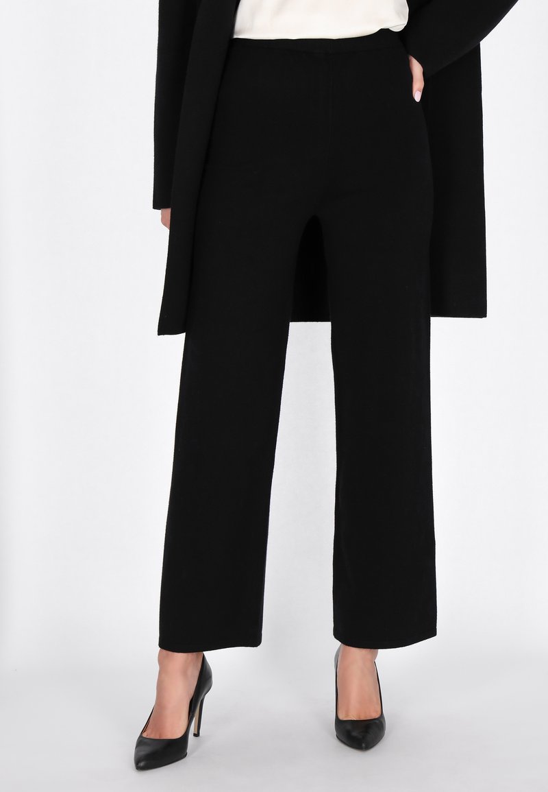 Zwarte, wijde broek met een gladde textuur en een hoge taille, gecombineerd met zwarte hoge hakken. Een minimalistische, moderne uitstraling.