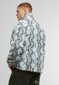 Fleece jakna s svetlo modrim in črnim vzorcem kačjega usnja, visokim ovratnikom, teksturirano blago in sproščenim krojem, v kombinaciji s temnimi hlačami.