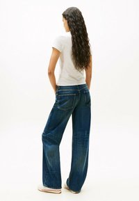 Jeans de mezclilla azul de pierna ancha con acabado deslavado, cinco bolsillos y cierre de botón. Vista trasera con una camiseta ajustada gris clara.