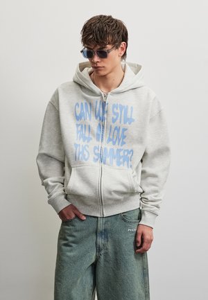 Mladý muž v sivom mikinovom pulóvri na zips s modrým nápisom „Môžeme sa tento rok v lete ešte zaľúbiť?“ a voľných modrých džínsoch, pózuje pred jednofarebným pozadím.