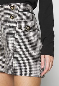 Élkové šortky s černobílým houndstooth vzorem, zdobené zlatými a černými knoflíky, nošené s černým topem s dlouhým rukávem.