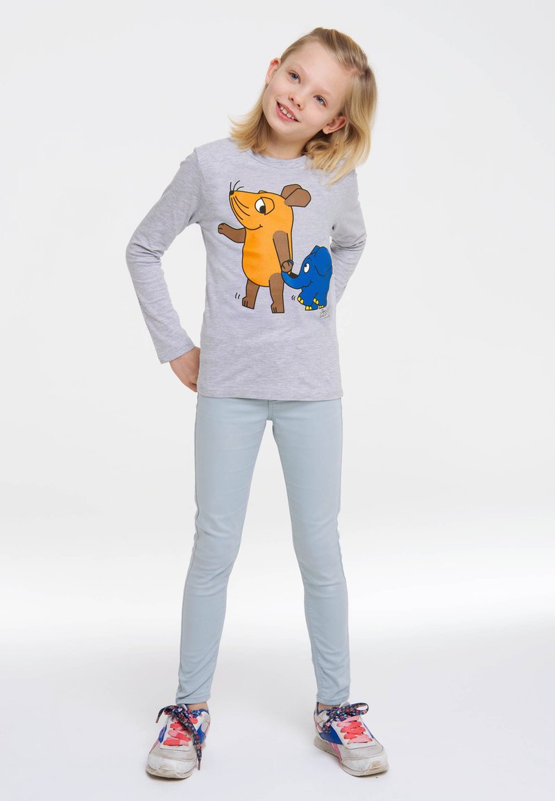 Graues langärmeliges Shirt mit einem orangefarbenen und braunen Cartoon-Tier-Print auf der Vorderseite, kombiniert mit hellblauen engen Hosen und bunten Turnschuhen.