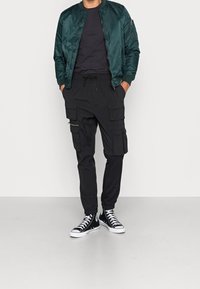 Tummanvihreä bomber-takki, yhdistetty mustiin taskullisiin cargo-housuihin. Malli käyttää mustia high-top tennareita.