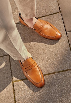 Mocassins en cuir beige portés avec un pantalon clair, debout sur un sol en pierre à l'extérieur.