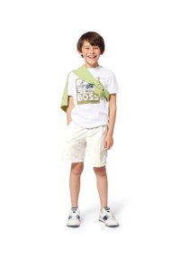 Maglietta bianca con una grafica dorata che reca la scritta "BOSS", maglione verde chiaro annodato attorno al collo, pantaloni corti leggeri a fantasie e scarpe da ginnastica bianche.