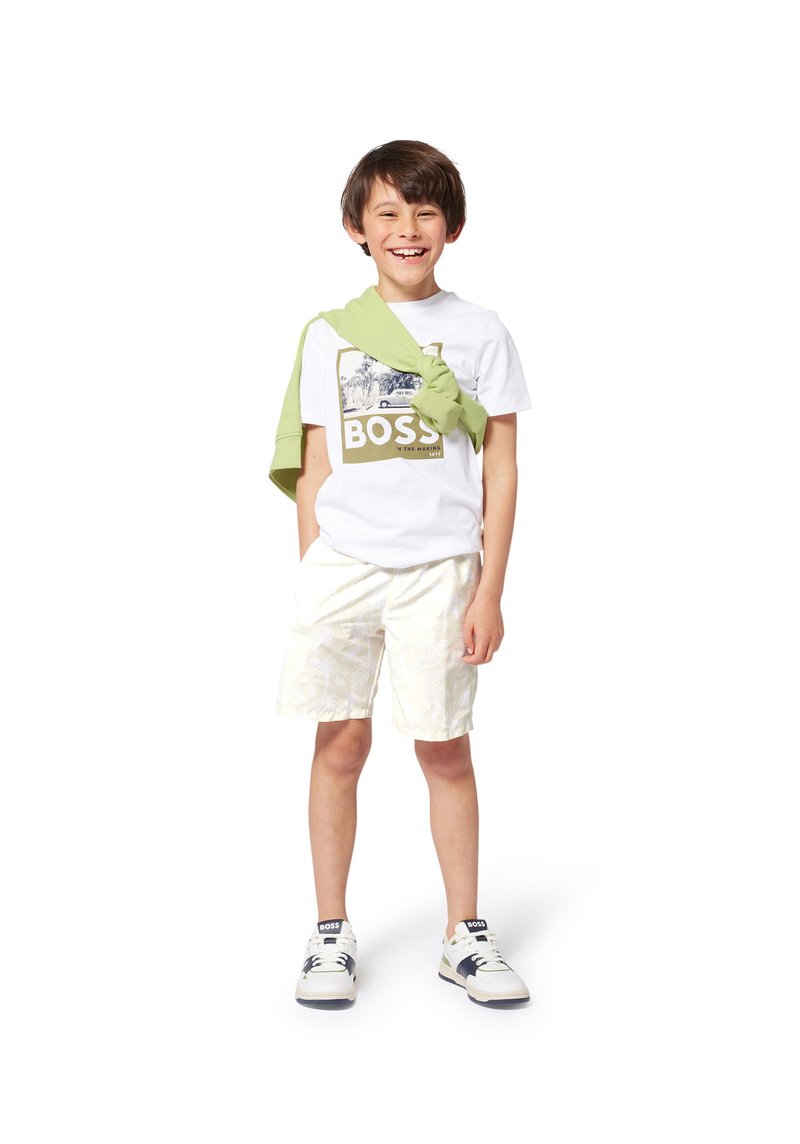 Maglietta bianca con una grafica dorata che reca la scritta "BOSS", maglione verde chiaro annodato attorno al collo, pantaloni corti leggeri a fantasie e scarpe da ginnastica bianche.