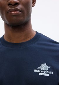 Tamsiai mėlynas sportinis megztinis su šonkačių apykakle. Krūtinėje yra baltas logotipo dizainas, kuriame daugiausia užrašyta "Marc O'Polo DENIM."