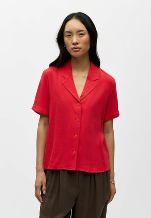 Femme aux longs cheveux noirs portant une chemise rouge à manches courtes boutonnée et un pantalon ample brun foncé, debout devant un fond blanc uni.