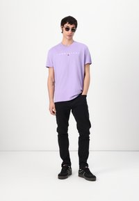 Lila Baumwoll-T-Shirt mit "TOMMY JEANS" Logo auf der Brust, kombiniert mit schwarzen Hosen und schwarzen Sneakers. Accessorized mit runden Sonnenbrillen.
