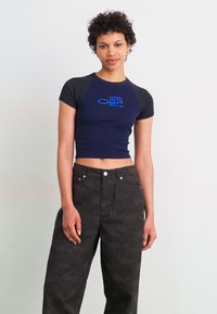 Marineblå crop top med korte sorte ærmer, med blå tekst, der er parret med høj talje grå bukser med et subtilt mønster og to forlommer.