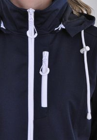 Marineblaue Softshell-Jacke mit hohem Kragen, weißen Reißverschlüssen, verstellbaren Kordelzügen und einem Mesh-Einsatz an der Kapuze für Belüftung.