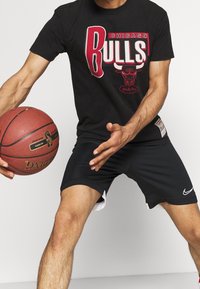 Zwarte katoenen T-shirt met rood en wit "Chicago Bulls" grafiek; zwarte sportbroek met witte accenten en Nike-logo; een basketbal vasthoudend.