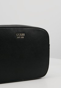 Schwarze, strukturierte Tasche mit goldfarbenem "GUESS EST 1981"-Logo. Sie hat eine rechteckige Form und glatte Akzente, die für verschiedene Anwendungen geeignet sind.