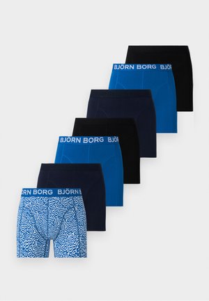 Modèle masculin portant un boxer Björn Borg bleu, assis de côté sur une chaise en métal avec assise beige, sur un fond clair uni.