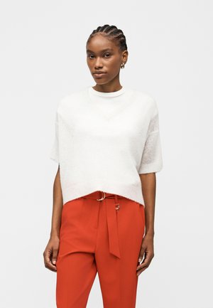 Młoda kobieta z warkoczami, nosząca złote kolczyki koła, luźny biały sweter z krótkim rękawem oraz wysokie czerwone spodnie z paskiem.