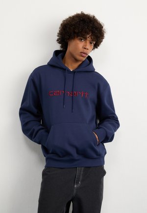 Sudadera azul marino hecha de algodón con un bolsillo frontal, que presenta un logotipo bordado en rojo de "Carhartt" y cordones ajustables en la capucha.