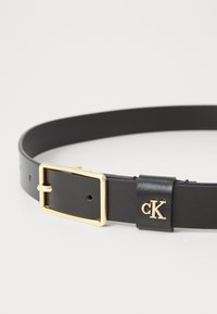 Zwarte leren riem met een gouden metalen gesp en een zwarte leren accent met het merk "cK." Glad textuur en verstelbare pasvorm.
